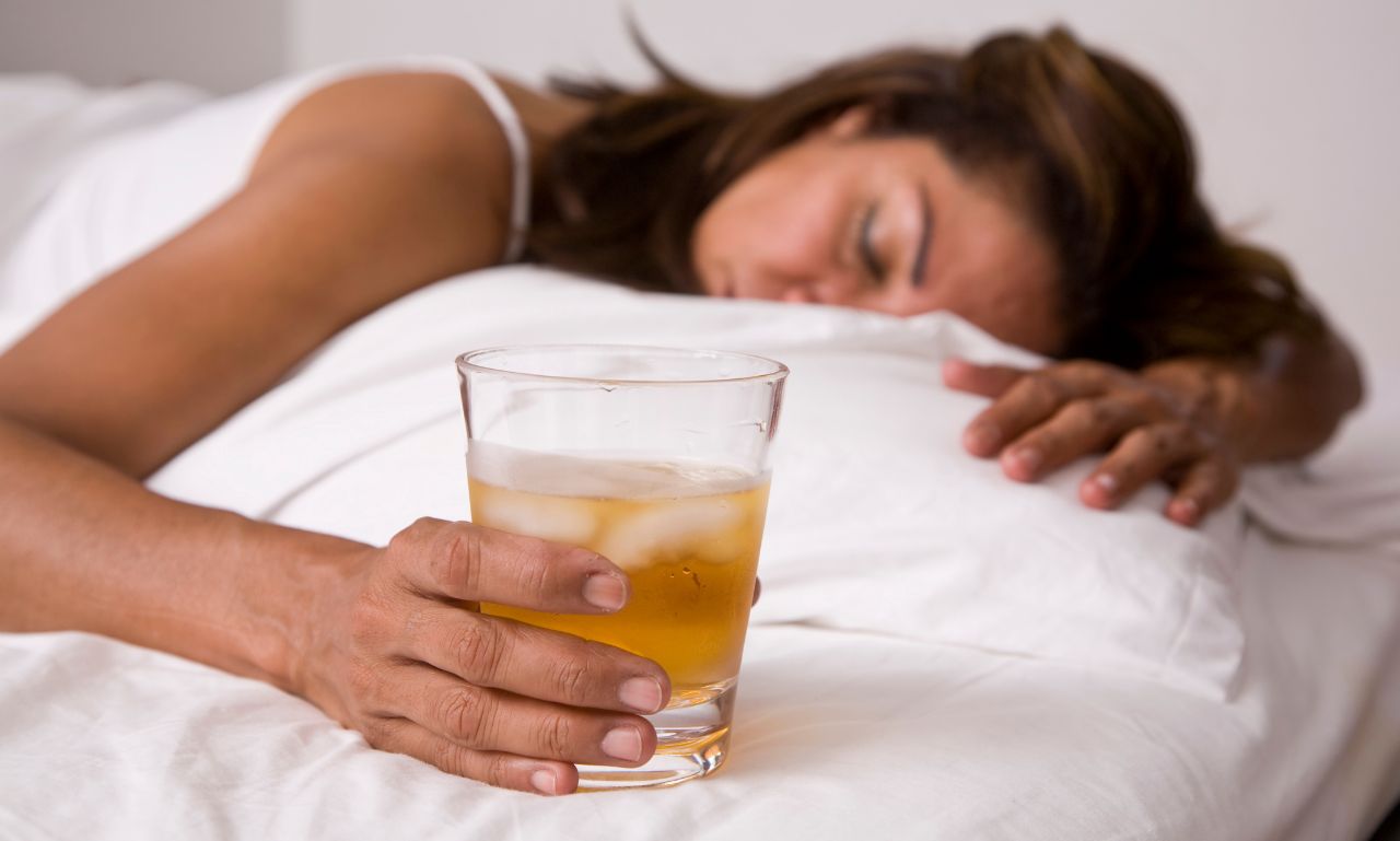 A woman lying in bed holding a glass of alcohol, symbolizing the complex risks and effects of alcohol consumption in women, highlighting gender-specific health concerns.