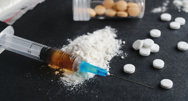A syringe with brown liquid, white powder, and scattered pills on a dark surface—depicting drug use paraphernalia linked to erratic or risky behavior associated with meth addiction.