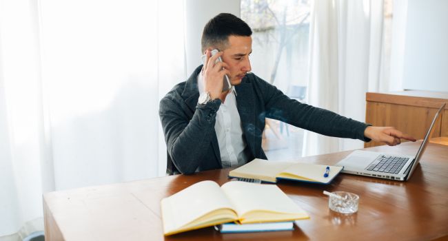 Man multitasking on phone and laptop with notebooks, illustrating balancing work responsibilities with outpatient treatment.