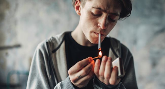 A young man with pale skin and a gaunt face lights a cigarette, illustrating the visible toll of meth addiction on physical appearance and health.