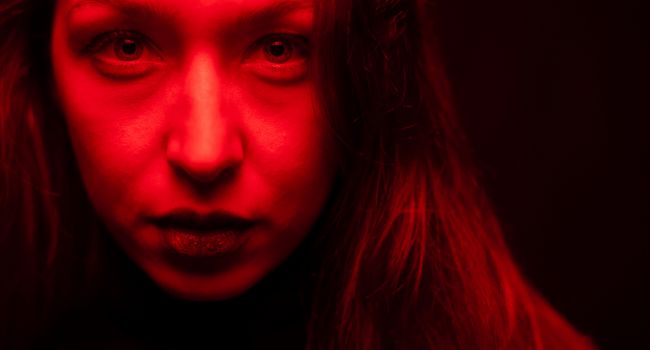 Close-up of a woman's face under red lighting, highlighting intense eyes and facial tension as potential physical signs of meth use.