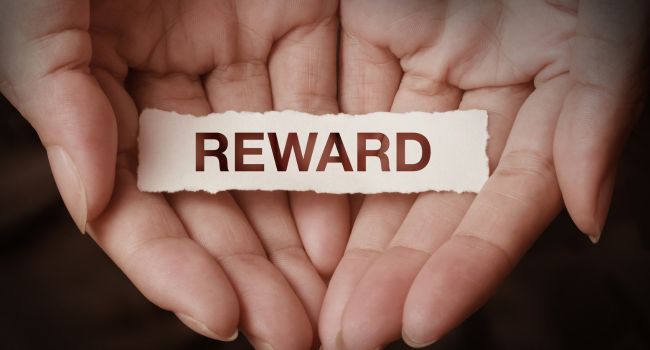 Open hands holding a piece of paper labeled “REWARD,” symbolizing the importance of designing an effective reward system to motivate progress in addiction recovery.