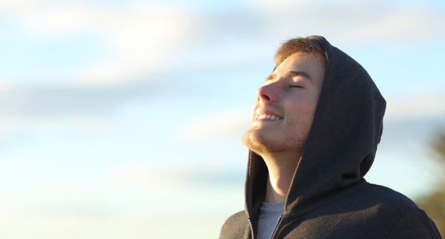 A young man wearing a hoodie, eyes closed and smiling toward the sky, illustrating calm awareness central to mindfulness practice.