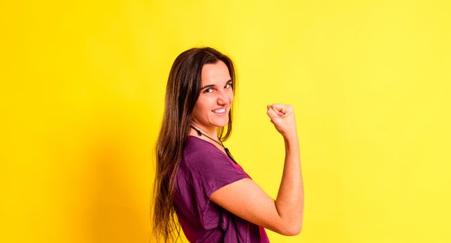 Confident woman smiling and flexing her arm against a yellow background, symbolizing self-esteem in meth addiction recovery.