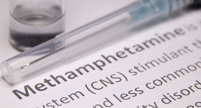 Close-up of a syringe and a document highlighting "Methamphetamine" and its central nervous system stimulant effects.