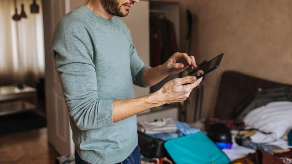 Man in a casual green sweater checking his tablet while packing a suitcase in a bedroom, preparing for a rehab stay.
