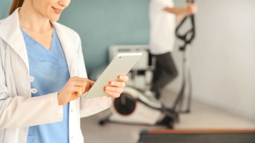 Smiling female healthcare professional using a digital tablet, with a blurred patient exercising in the background during outpatient care.