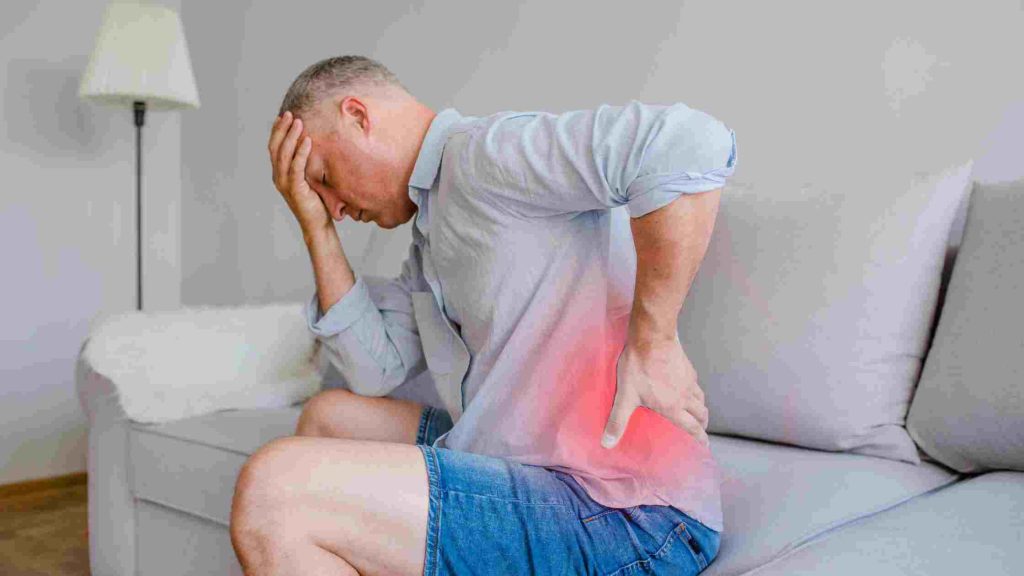 Man sitting on sofa, holding his lower back and forehead in discomfort, red highlight showing pain area.
