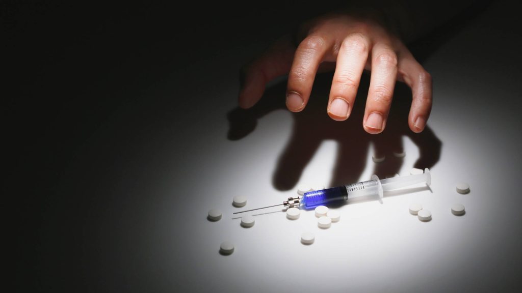 A hand reaching toward a syringe and pills in dim light, symbolizing addiction and FMLA coverage for drug and alcohol rehab.